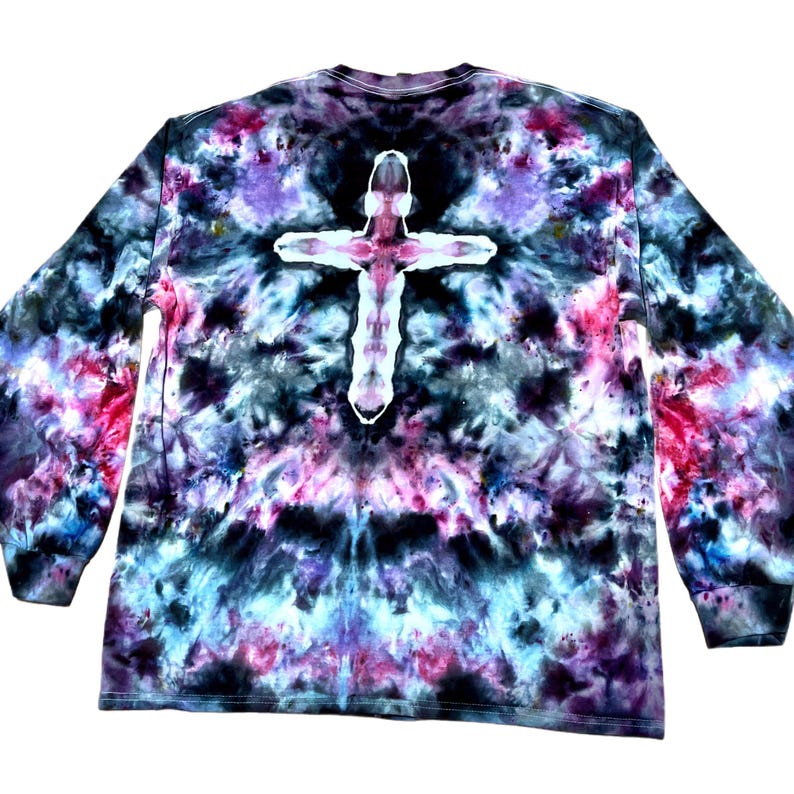 May include: A long-sleeve shirt featuring a tie-dye pattern in shades of black, purple, blue, and pink. The center of the garment displays a prominent white cross shape created through the tie-dye process, set against the dark, mottled background.