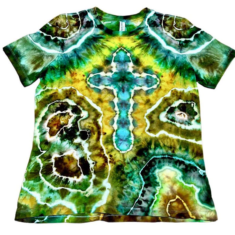 Puede incluir: Una camiseta de manga corta te&ntilde;ida con tinte en tonos verdes, amarillos y azules. La camiseta presenta un dise&ntilde;o de cruz central y un patr&oacute;n abstracto. La tela parece suave y c&oacute;moda.