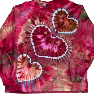 Może przedstawiać: Koszulka z długim rękawem i okrągłym dekoltem z wzorem tie-dye w odcieniach czerwieni, r&oacute;żu i brązu. Odzież posiada trzy wyraźne motywy w kształcie serca z białym obrysem, rozmieszczone ukośnie na przedniej części ubrania.