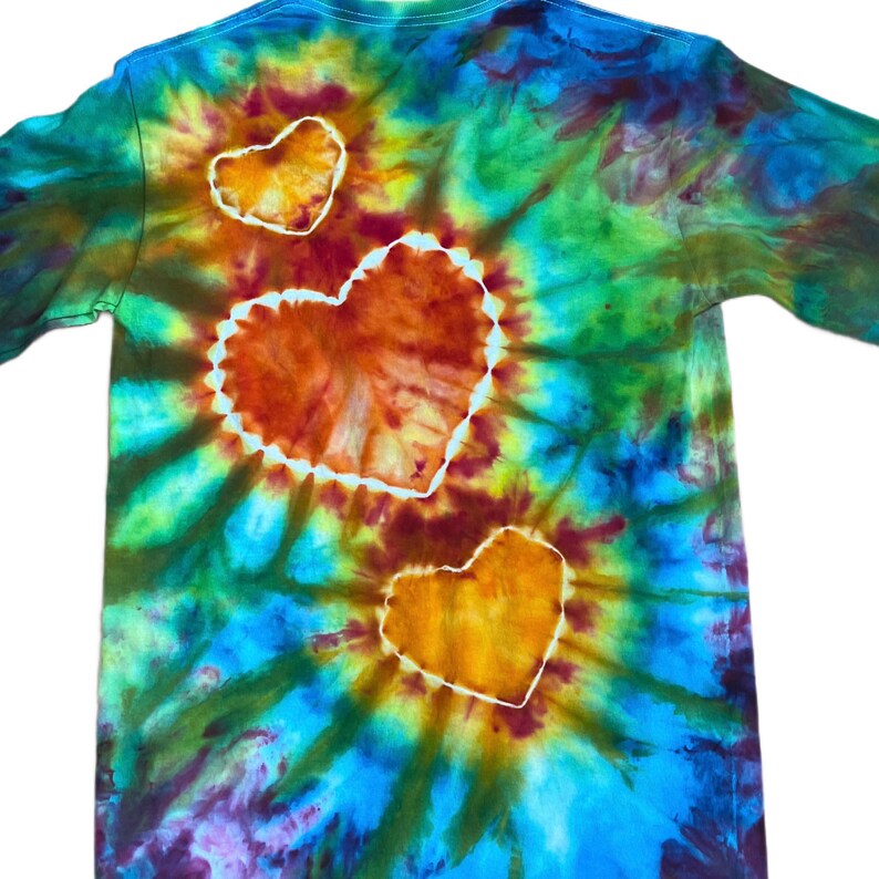 Może przedstawiać: Koszulka z długim rękawem z żywym wzorem tie-dye w odcieniach niebieskiego, zielonego, ż&oacute;łtego i pomarańczowego. Wz&oacute;r zawiera trzy wyraźne kształty serc z białym obrysem, w tym jedno duże serce centralne i dwa mniejsze serca.