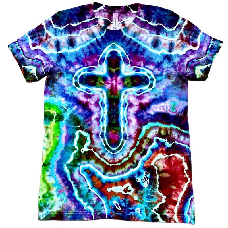 May include: A tie-dye t-shirt with a cross design. The shirt features a vibrant mix of colours, including blue, purple, green, and red. The cross is centered on the front, with a white outline. The shirt is made of a soft material.
