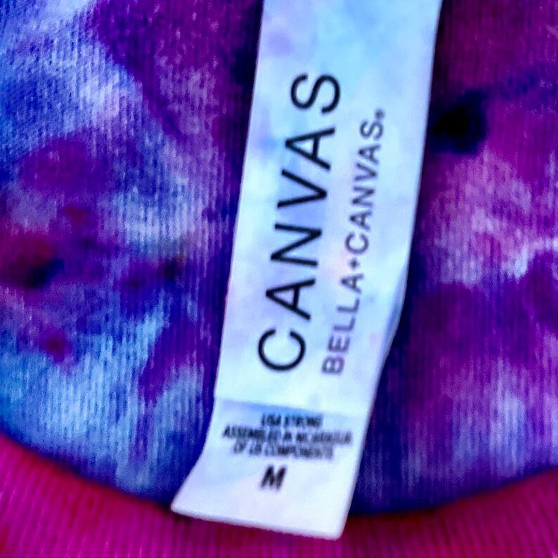 May include: A close-up of a white tag with the word "CANVAS" in large, bold, black letters. Below it, the text "BELLA+CANVAS." is visible. The tag also includes additional text and the letter "M". The background is a vibrant mix of pink, purple, and blue.