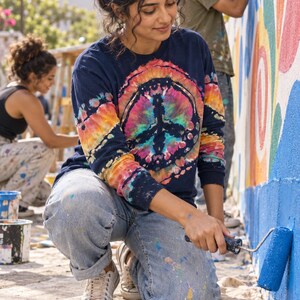 May include: A long-sleeve navy blue shirt featuring a vibrant, multi-colored tie-dye peace sign graphic on the front. The shirt has a crew neckline and is worn with light-wash denim jeans, showcasing a casual style for artistic activities.