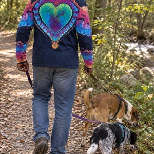 May include: A long-sleeve navy blue shirt featuring a large, vibrant tie-dye heart design on the back in shades of teal, purple, and pink, paired with blue denim jeans and brown hiking boots, while walking two dogs on leashes along a trail.
