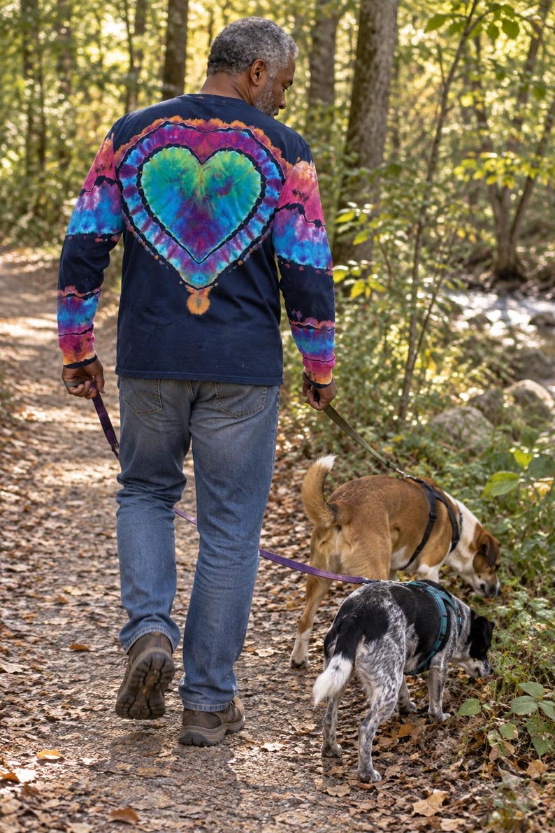 May include: A long-sleeve navy blue shirt featuring a large, vibrant tie-dye heart design on the back in shades of teal, purple, and pink, paired with blue denim jeans and brown hiking boots, while walking two dogs on leashes along a trail.