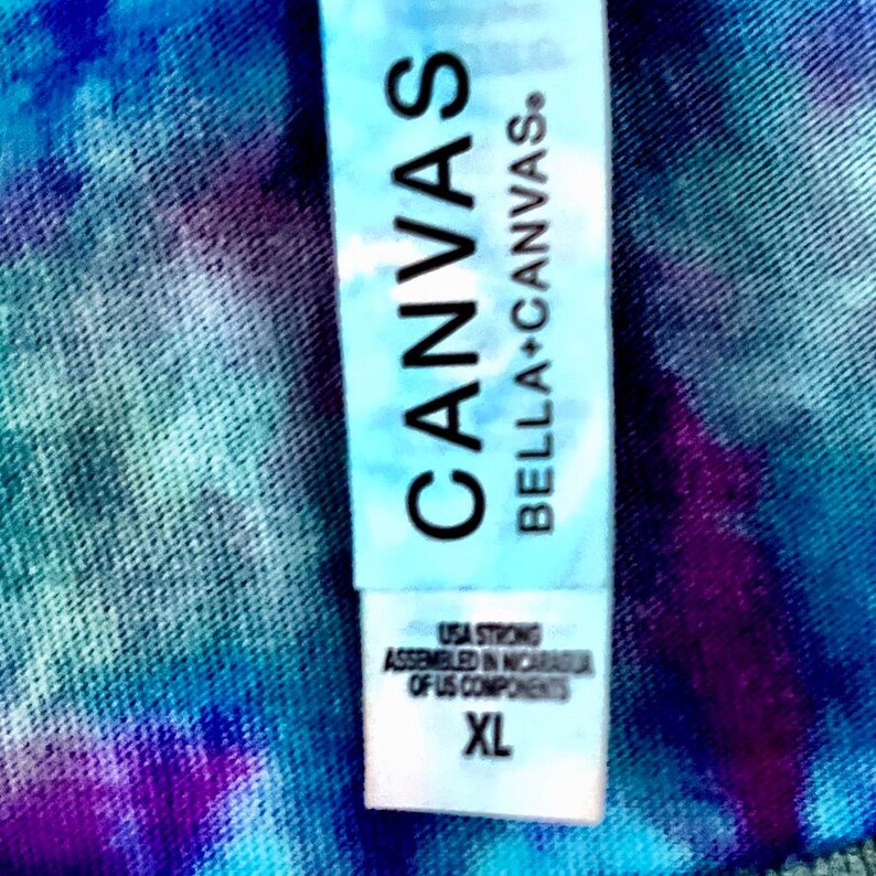 Op de afbeelding: Close-up van een tie-dye shirt met een label met de tekst "CANVAS BELLA+CANVAS". Het label vermeldt ook "USA STRONG ASSEMBLED IN NICARAGUA OF US COMPONENTS" en de maat "XL".