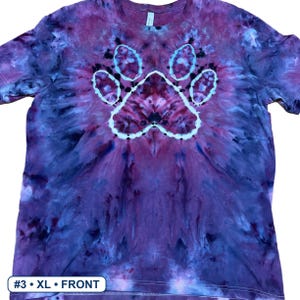 Może przedstawiać: Koszulka z kr&oacute;tkim rękawem z wzorem tie-dye w odcieniach fioletu, błękitu i czerni. Na środku koszulki znajduje się wyraźna biała grafika przedstawiająca odcisk łapy. Mała biała etykieta w lewym dolnym rogu zawiera napis #3 &bull; XL &bull; FRONT.