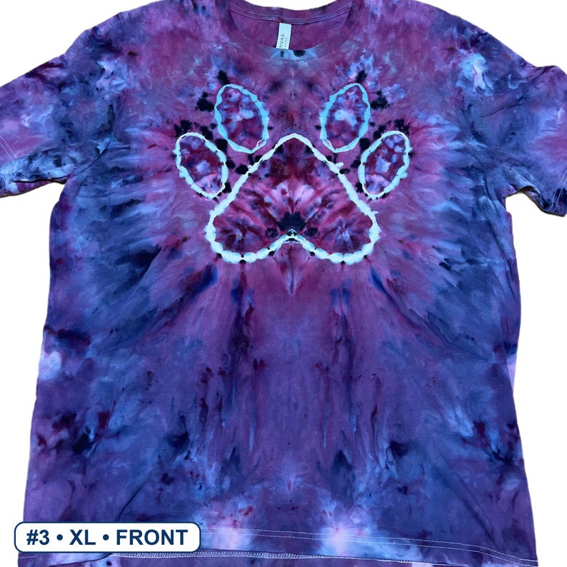 Może przedstawiać: Koszulka z kr&oacute;tkim rękawem z wzorem tie-dye w odcieniach fioletu, błękitu i czerni. Na środku koszulki znajduje się wyraźna biała grafika przedstawiająca odcisk łapy. Mała biała etykieta w lewym dolnym rogu zawiera napis #3 &bull; XL &bull; FRONT.