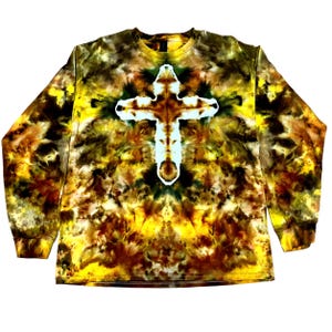 Puede incluir: Una camiseta de manga larga con cuello redondo que presenta un patr&oacute;n tie-dye en tonos amarillo, marr&oacute;n y negro. El centro de la prenda muestra una prominente forma de cruz blanca creada mediante el proceso de te&ntilde;ido sobre el fondo jaspeado.