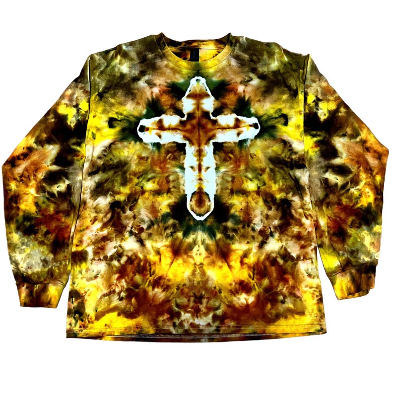 Puede incluir: Una camiseta de manga larga con cuello redondo que presenta un patr&oacute;n tie-dye en tonos amarillo, marr&oacute;n y negro. El centro de la prenda muestra una prominente forma de cruz blanca creada mediante el proceso de te&ntilde;ido sobre el fondo jaspeado.