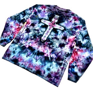 May include: A long-sleeve shirt featuring a tie-dye pattern in shades of black, purple, blue, and pink. The center of the shirt displays a prominent white cross shape that is void of the tie-dye color, creating a stark contrast against the dark background.