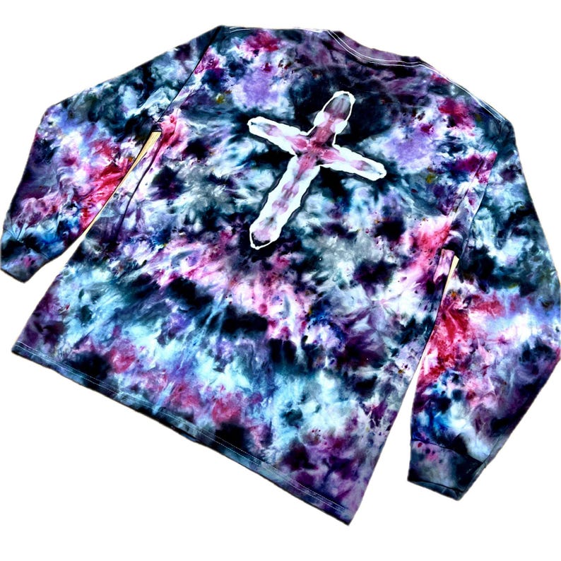 May include: A long-sleeve shirt featuring a tie-dye pattern in shades of black, purple, blue, and pink. The center of the shirt displays a prominent white cross shape that is void of the tie-dye color, creating a stark contrast against the dark background.