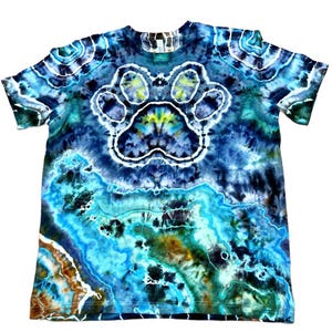 May include: A tie-dye t-shirt with a large paw print design in the center. The shirt features a blend of blues, greens, and browns, creating a unique, marbled effect. The paw print is outlined in white.