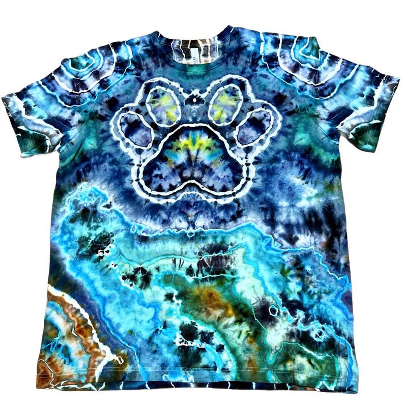 May include: A tie-dye t-shirt with a large paw print design in the center. The shirt features a blend of blues, greens, and browns, creating a unique, marbled effect. The paw print is outlined in white.