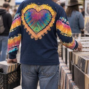 May include: A navy blue long-sleeve shirt featuring a large, central tie-dye heart design on the back in vibrant rainbow colors. The sleeves also display matching tie-dye patterns in orange, yellow, and blue stripes against the dark fabric.