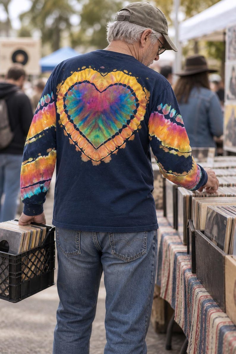 May include: A navy blue long-sleeve shirt featuring a large, central tie-dye heart design on the back in vibrant rainbow colors. The sleeves also display matching tie-dye patterns in orange, yellow, and blue stripes against the dark fabric.