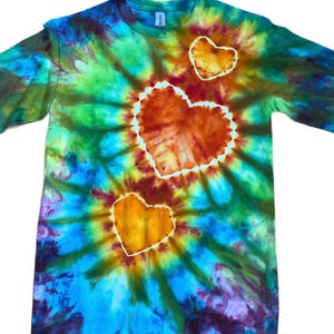 Może przedstawiać: Koszulka z kr&oacute;tkim rękawem i okrągłym dekoltem, ozdobiona żywym wzorem tie-dye w odcieniach niebieskiego, zielonego i ż&oacute;łtego. Z przodu znajdują się trzy wyraźne motywy w kształcie serca w kolorze pomarańczowym i ż&oacute;łtym z białym obramowaniem.