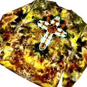 Puede incluir: Una camiseta de manga larga con cuello redondo que presenta un patr&oacute;n de te&ntilde;ido anudado en tonos amarillo, marr&oacute;n y verde oscuro. El centro de la prenda muestra una prominente forma de cruz blanca creada mediante una t&eacute;cnica de te&ntilde;ido inverso.