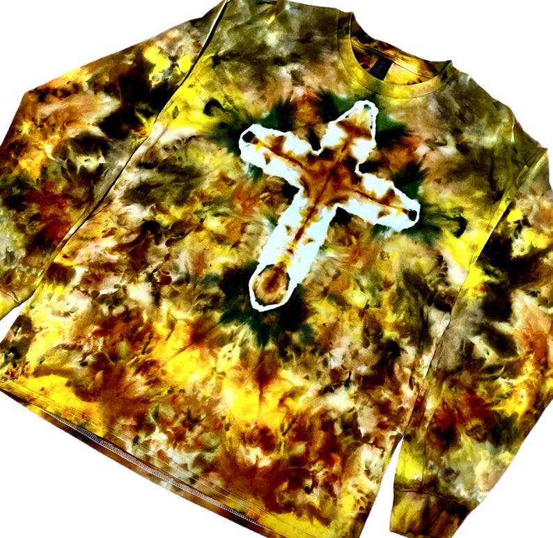 Puede incluir: Una camiseta de manga larga con cuello redondo que presenta un patr&oacute;n de te&ntilde;ido anudado en tonos amarillo, marr&oacute;n y verde oscuro. El centro de la prenda muestra una prominente forma de cruz blanca creada mediante una t&eacute;cnica de te&ntilde;ido inverso.