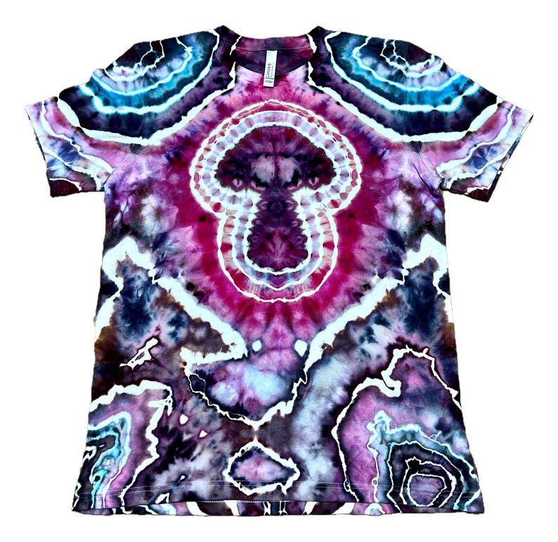 Puede incluir: Una camiseta de manga corta con un complejo patr&oacute;n de te&ntilde;ido anudado en tonos p&uacute;rpura intenso, magenta, negro y cian brillante. El dise&ntilde;o muestra formas circulares conc&eacute;ntricas similares a geodas con contornos blancos sobre la tela.