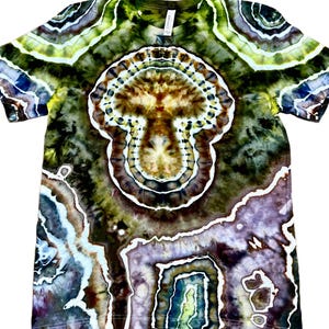 May include: A short-sleeve t-shirt featuring a complex tie-dye pattern in shades of olive green, brown, purple, and white. The design displays concentric circular and organic shapes that resemble a geode or mushroom-like central motif.