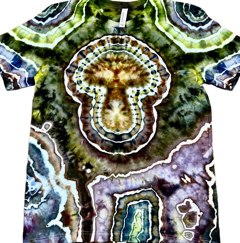 May include: A short-sleeve t-shirt featuring a complex tie-dye pattern in shades of olive green, brown, purple, and white. The design displays concentric circular and organic shapes that resemble a geode or mushroom-like central motif.