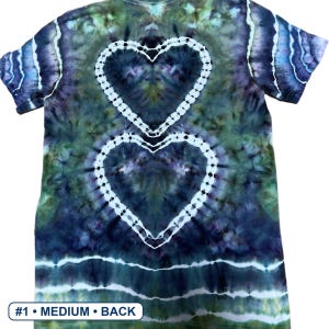Może przedstawiać: Koszulka z kr&oacute;tkim rękawem z wzorem tie-dye w odcieniach ciemnego błękitu, leśnej zieleni i fioletu. Wz&oacute;r zawiera dwa ułożone jeden na drugim białe kontury serc na środku plec&oacute;w oraz poziome białe paski przy dolnej krawędzi. Tekst #1 MEDIUM BACK.