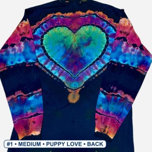 May include: A long-sleeve shirt with a black base featuring a tie-dye heart design in shades of blue, green, purple, and orange. The sleeves have horizontal tie-dye stripes. Text at the bottom reads #1 &bull; MEDIUM &bull; PUPPY LOVE &bull; BACK.