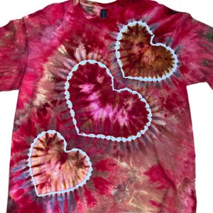 Może przedstawiać: Koszulka z kr&oacute;tkim rękawem z wzorem tie-dye w odcieniach czerwieni, r&oacute;żu i ciemnego brązu. Na koszulce znajdują się trzy wyraźne motywy w kształcie serca, każdy z nich obwiedziony wyraźną białą linią, kt&oacute;ra kontrastuje z żywymi, wirującymi kolorami.