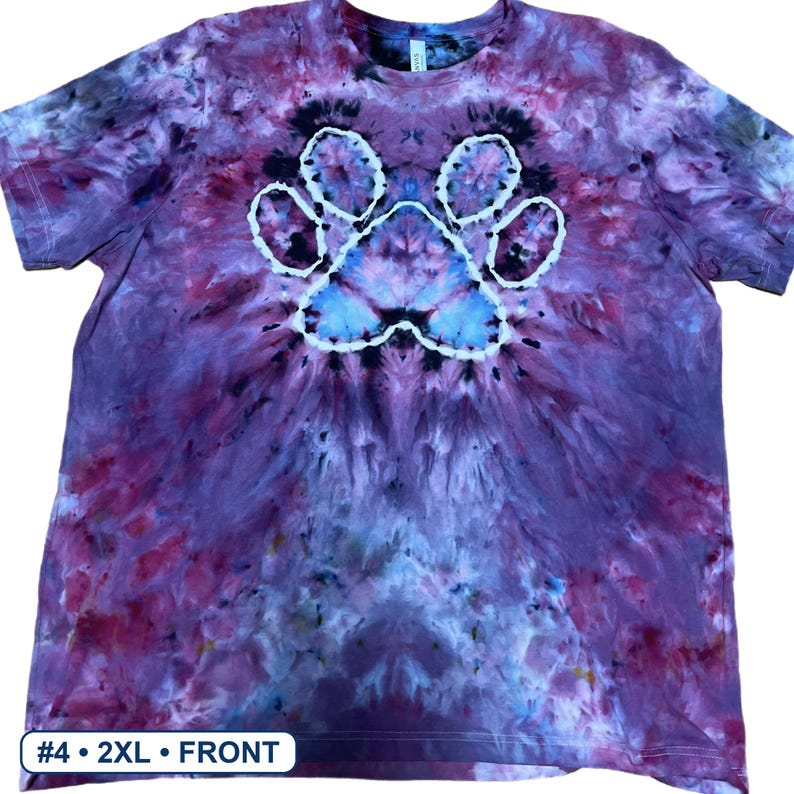 Może przedstawiać: Koszulka z kr&oacute;tkim rękawem z wzorem tie-dye w odcieniach fioletu, błękitu i czerni. Na środku koszulki znajduje się biały zarys odcisku łapy. W dolnym rogu znajduje się biała etykieta z napisem #4 &bull; 2XL &bull; FRONT.