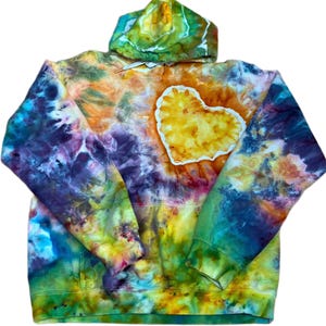 May include: A long-sleeve hooded sweatshirt featuring a vibrant tie-dye pattern in shades of yellow, orange, purple, blue, and green. A prominent yellow heart-shaped patch with a white border is centered on the chest area of the garment.