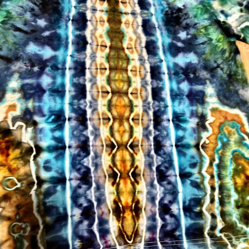 May include: A close-up of a tie-dye textile with a vibrant pattern. The design features vertical stripes in shades of blue, turquoise, yellow, and brown. The fabric has a soft, textured appearance, with a unique, artistic aesthetic.