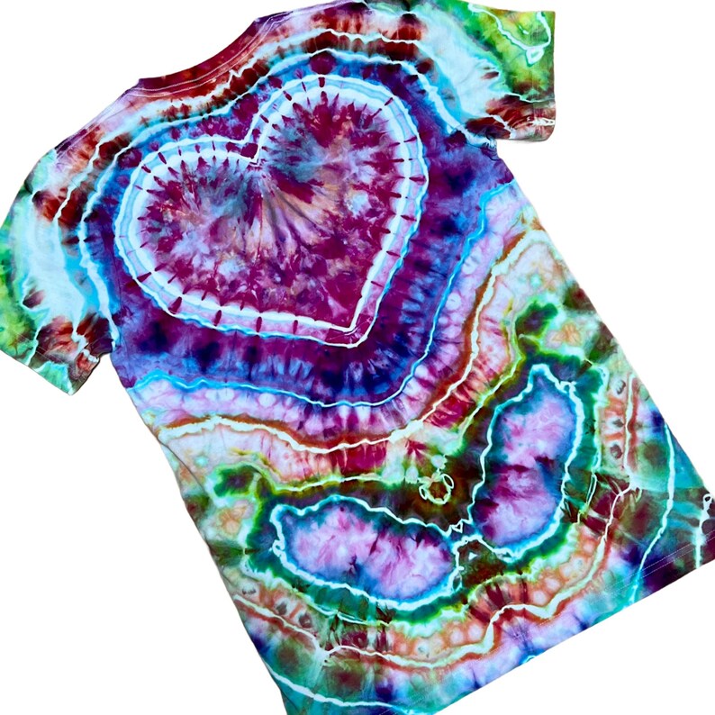 May include: A short-sleeve t-shirt featuring a tie-dye pattern with a prominent central heart shape. The design incorporates vibrant shades of magenta, purple, teal, green, and white, creating a swirling, multi-layered effect across the fabric.