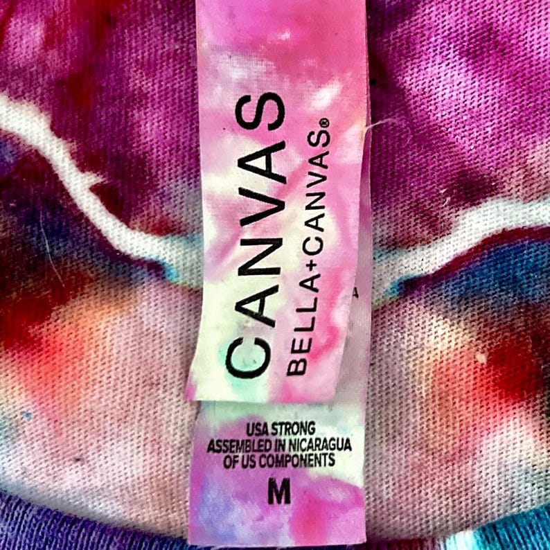 May include: Close-up of a tie-dye shirt tag. The tag is white and pink with the word "CANVAS" in large black letters. Below the word "CANVAS" is "BELLA+CANVAS&reg;". The tag also reads "USA STRONG ASSEMBLED IN NICARAGUA OF US COMPONENTS" and has an "M" for size.
