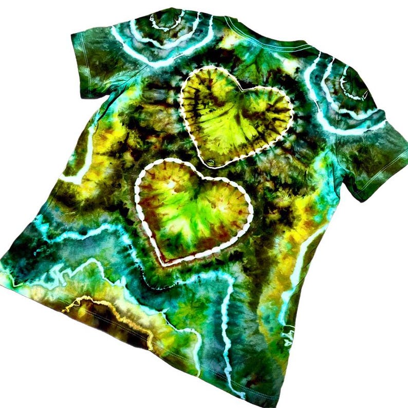 Puede incluir: Una camiseta te&ntilde;ida con tinte en tonos verdes, amarillos y azules. Dos grandes formas de coraz&oacute;n est&aacute;n delineadas en blanco en la parte posterior de la camiseta. La camiseta tiene mangas cortas y cuello redondo.