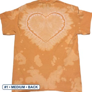 Peut inclure: Un t-shirt orange &agrave; manches courtes pr&eacute;sentant un motif tie-dye d&eacute;color&eacute; avec une forme de c&oelig;ur pro&eacute;minente centr&eacute;e au dos. Le tissu affiche des taches claires irr&eacute;guli&egrave;res sur toute la surface. Une &eacute;tiquette rectangulaire indique #1 &bull; MEDIUM &bull; BACK.