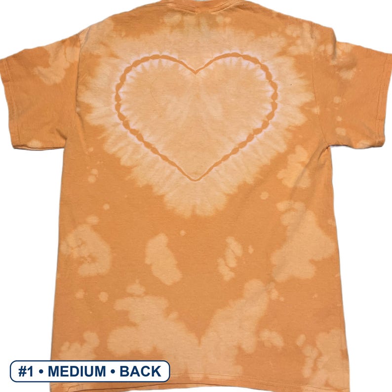 Peut inclure: Un t-shirt orange &agrave; manches courtes pr&eacute;sentant un motif tie-dye d&eacute;color&eacute; avec une forme de c&oelig;ur pro&eacute;minente centr&eacute;e au dos. Le tissu affiche des taches claires irr&eacute;guli&egrave;res sur toute la surface. Une &eacute;tiquette rectangulaire indique #1 &bull; MEDIUM &bull; BACK.