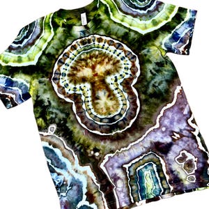 May include: A short-sleeve crew neck t-shirt featuring a complex tie-dye pattern with concentric, geode-like shapes in shades of olive green, brown, deep purple, and white. The design covers the entire garment, creating a layered, organic aesthetic.