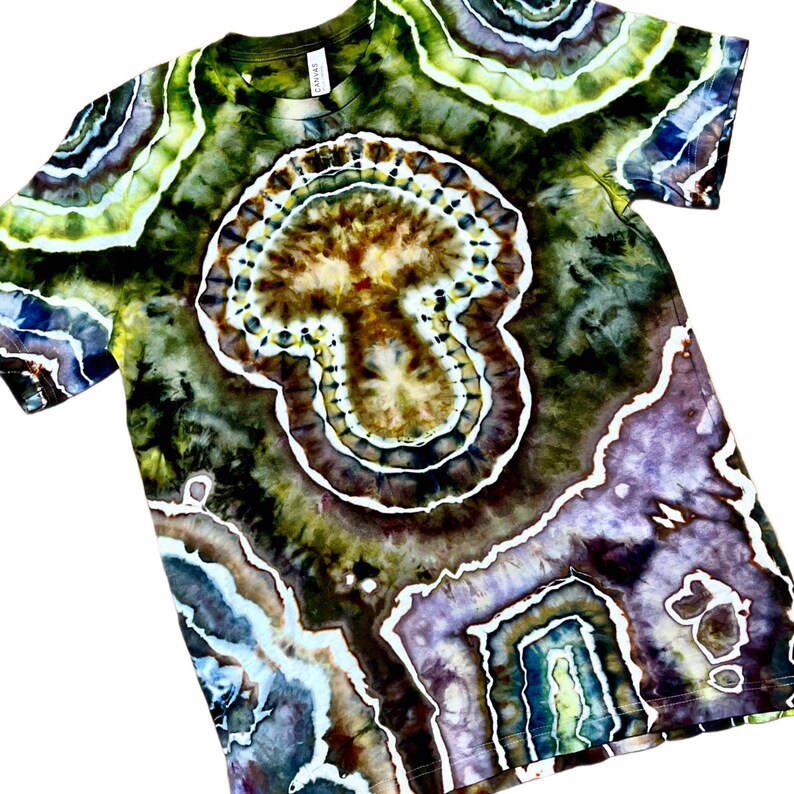 May include: A short-sleeve crew neck t-shirt featuring a complex tie-dye pattern with concentric, geode-like shapes in shades of olive green, brown, deep purple, and white. The design covers the entire garment, creating a layered, organic aesthetic.