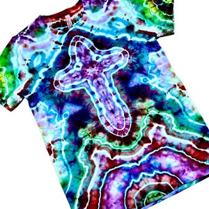 May include: A short-sleeve t-shirt featuring a vibrant tie-dye pattern in shades of blue, purple, green, and red. The design includes a prominent white outlined cross shape centered on the chest, surrounded by swirling, marbled color gradients.
