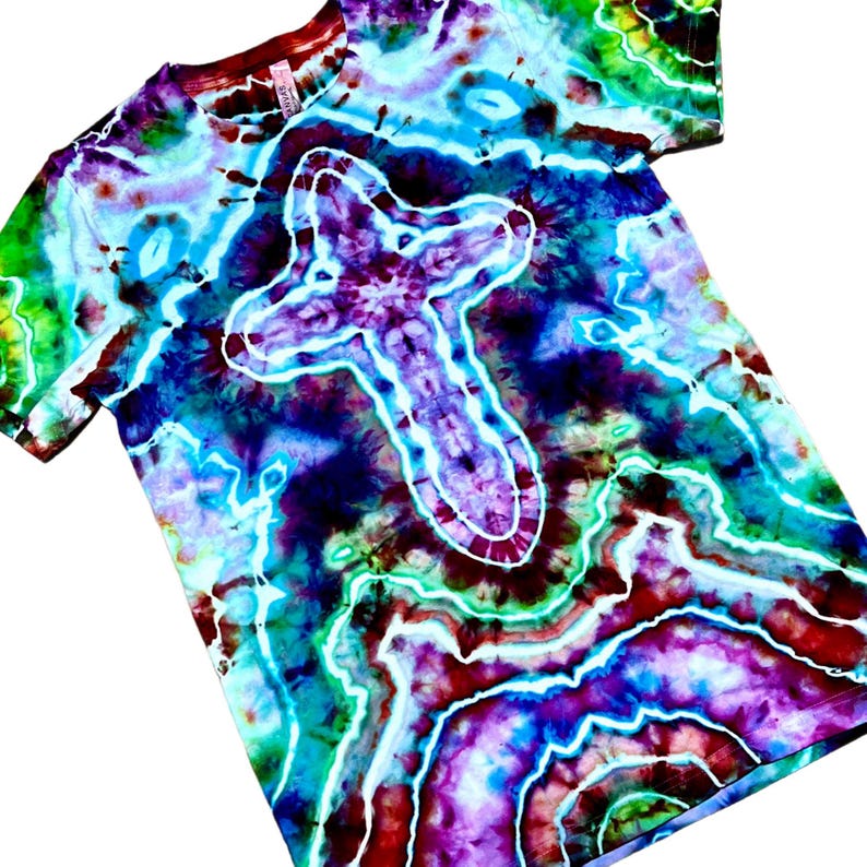 May include: A short-sleeve t-shirt featuring a vibrant tie-dye pattern in shades of blue, purple, green, and red. The design includes a prominent white outlined cross shape centered on the chest, surrounded by swirling, marbled color gradients.