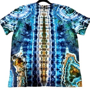 May include: A tie-dye t-shirt in shades of blue, green, and brown. The shirt has a symmetrical pattern with vertical stripes of color and swirling designs. The shirt is hanging on a black hanger.