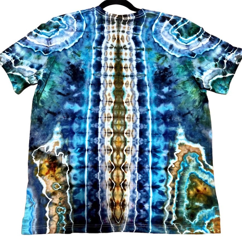 May include: A tie-dye t-shirt in shades of blue, green, and brown. The shirt has a symmetrical pattern with vertical stripes of color and swirling designs. The shirt is hanging on a black hanger.