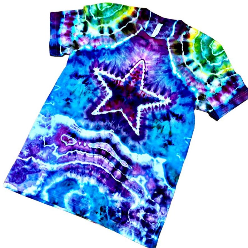 May include: A tie-dye t-shirt with a large star design. The shirt features a vibrant mix of colors, including blues, purples, and greens. The star is outlined in white, creating a striking contrast against the colorful background. The shirt is a short-sleeved crew neck.