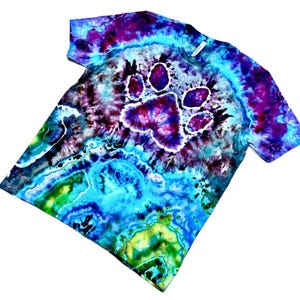 Op de afbeelding: Tie-dye T-shirt met een pootafdruk ontwerp. Het shirt heeft een levendige mix van kleuren, waaronder blauw, paars en groen. De pootafdruk staat centraal op de borst, wat zorgt voor een unieke en opvallende uitstraling.