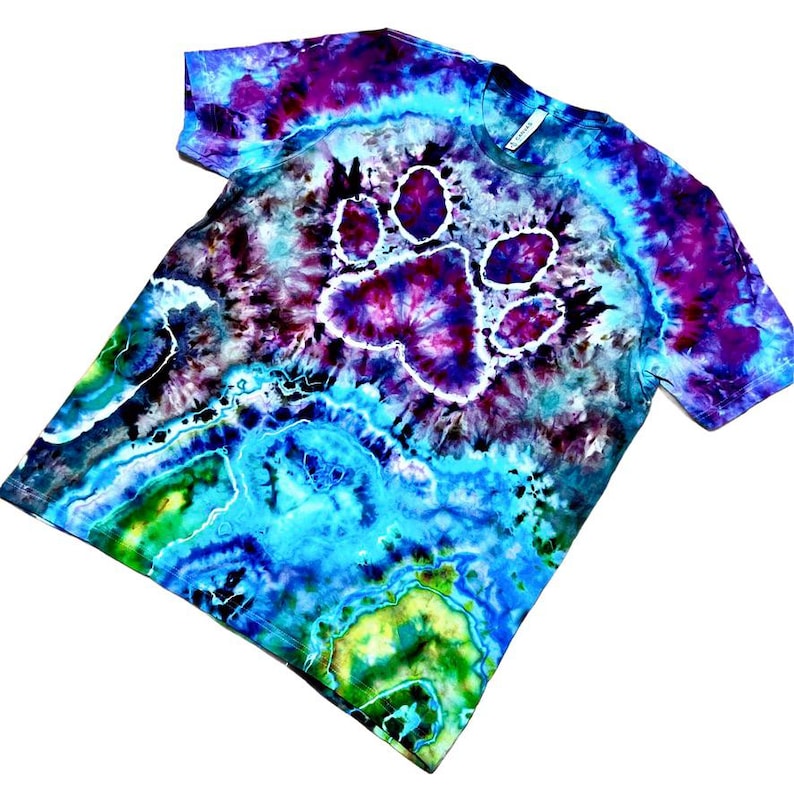 Op de afbeelding: Tie-dye T-shirt met een pootafdruk ontwerp. Het shirt heeft een levendige mix van kleuren, waaronder blauw, paars en groen. De pootafdruk staat centraal op de borst, wat zorgt voor een unieke en opvallende uitstraling.