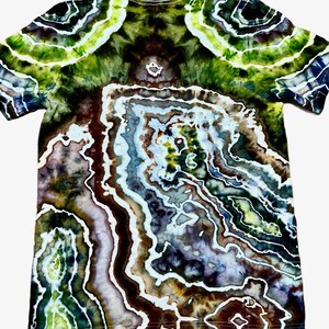 May include: A short-sleeve t-shirt featuring a complex tie-dye pattern that mimics the appearance of a geode or agate stone. The design incorporates swirling layers of deep forest green, dark brown, muted purple, and bright lime with white lines.