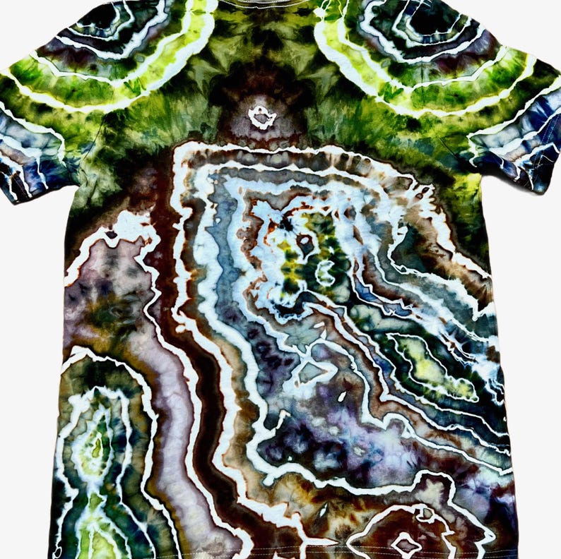 May include: A short-sleeve t-shirt featuring a complex tie-dye pattern that mimics the appearance of a geode or agate stone. The design incorporates swirling layers of deep forest green, dark brown, muted purple, and bright lime with white lines.