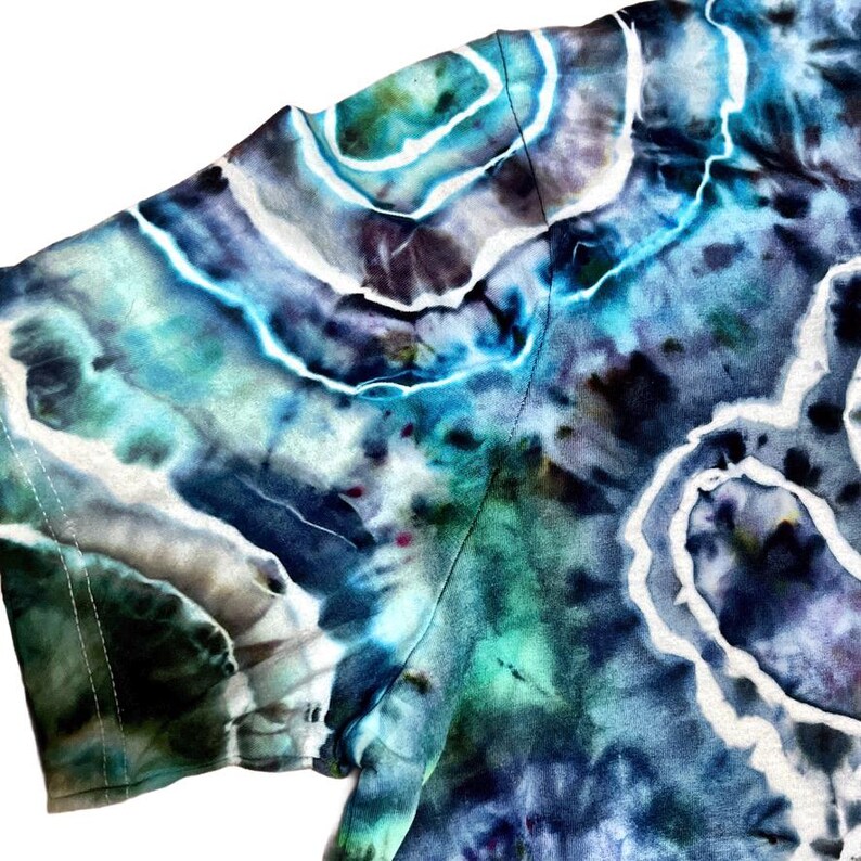 May include: Close-up of a tie-dye t-shirt in shades of blue, teal, and purple. The fabric has a swirling pattern with white accents, creating a unique and artistic design. The shirt's texture is visible, showcasing the dye's intricate details.