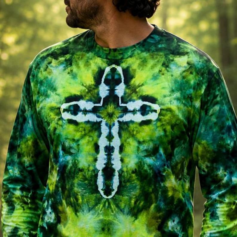 May include: A long-sleeve crew neck shirt featuring a tie-dye pattern in shades of forest green, lime green, and black. The center of the shirt displays a prominent white cross shape created through the tie-dye process, contrasting with the dark background.